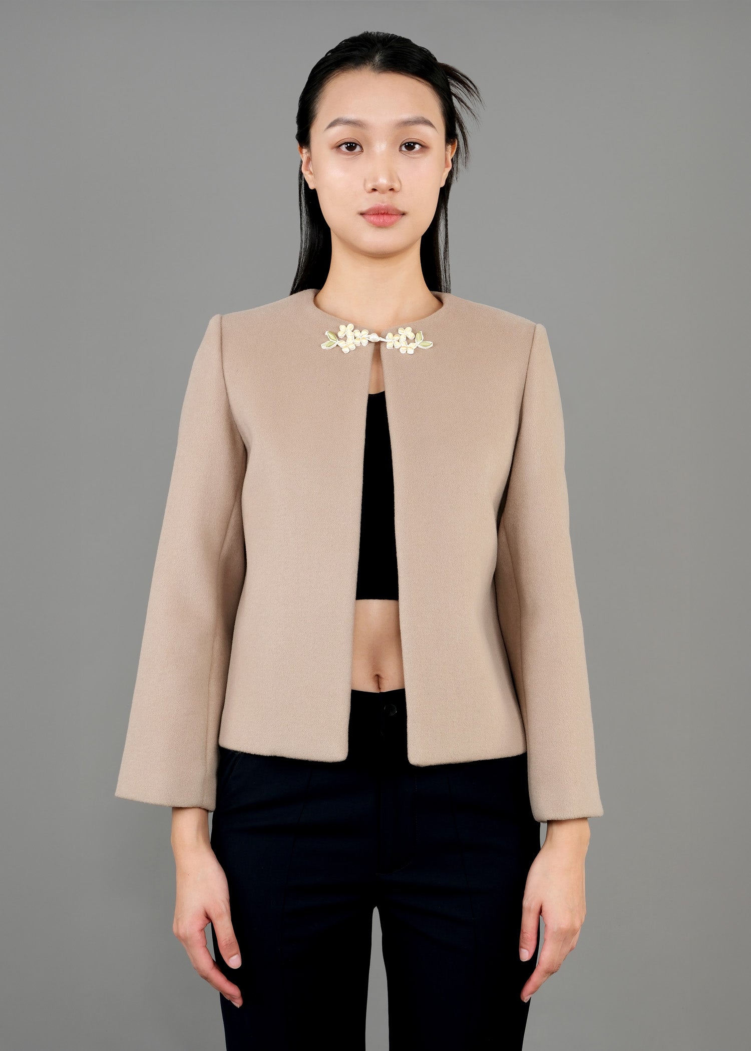 A woman stands in front view, wearing a stylish beige jacket with a round neckline and a decorative floral brooch at the front. The jacket has long sleeves and is paired with a black cropped top and dark trousers. 
一名女子站在正面，身穿時尚的米色圓領外套，胸前飾有花朵胸針。外套為長袖設計，內搭黑色露臍上衣和深色長褲。