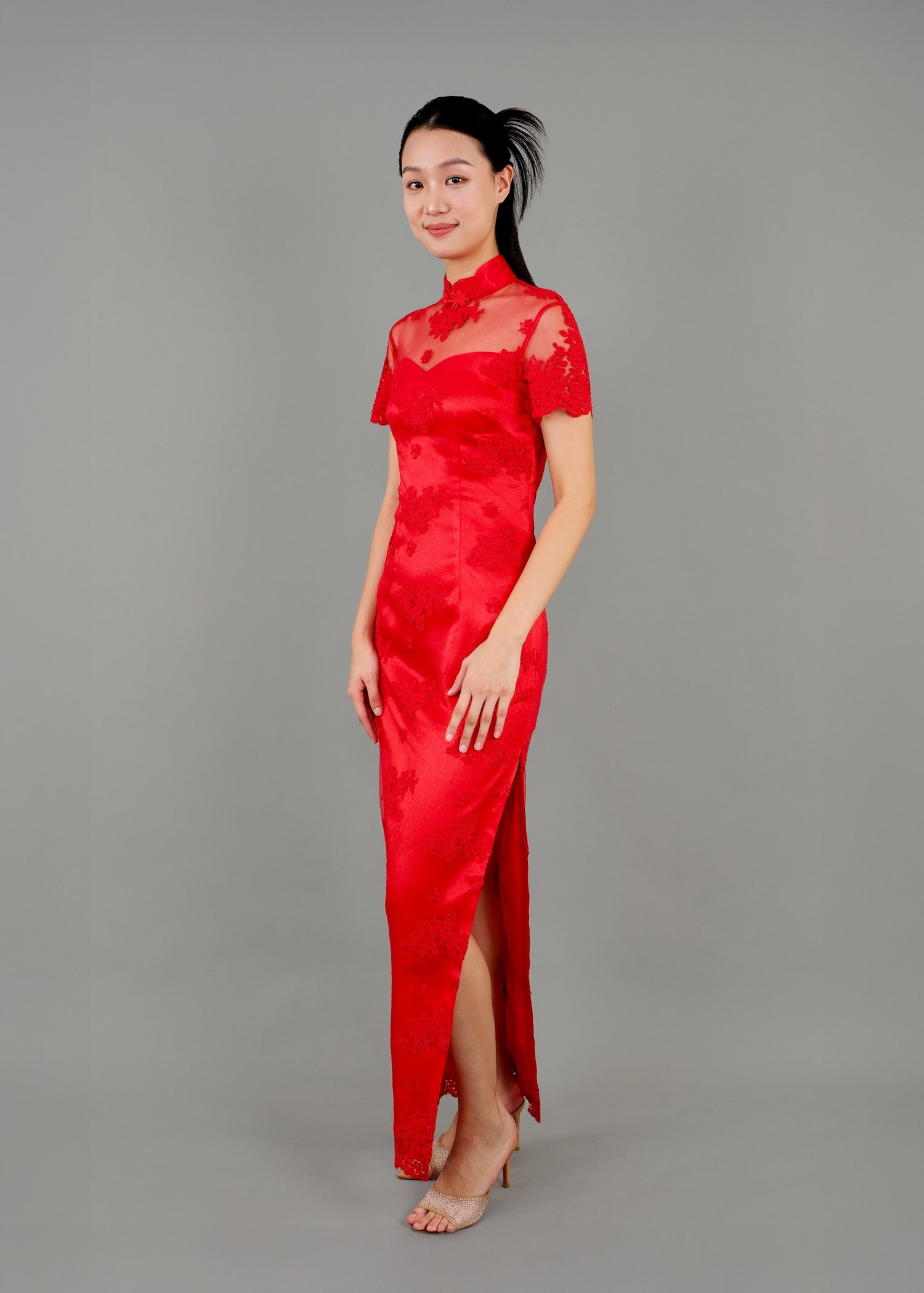 A woman stands in sidie view, wearing a red high-neck dress with sheer sleeves, floral lace details, and a side slit.
一名女子站在側面，身穿紅色高領連身裙，透明袖子上帶有花卉蕾絲細節，並有側開衩。


