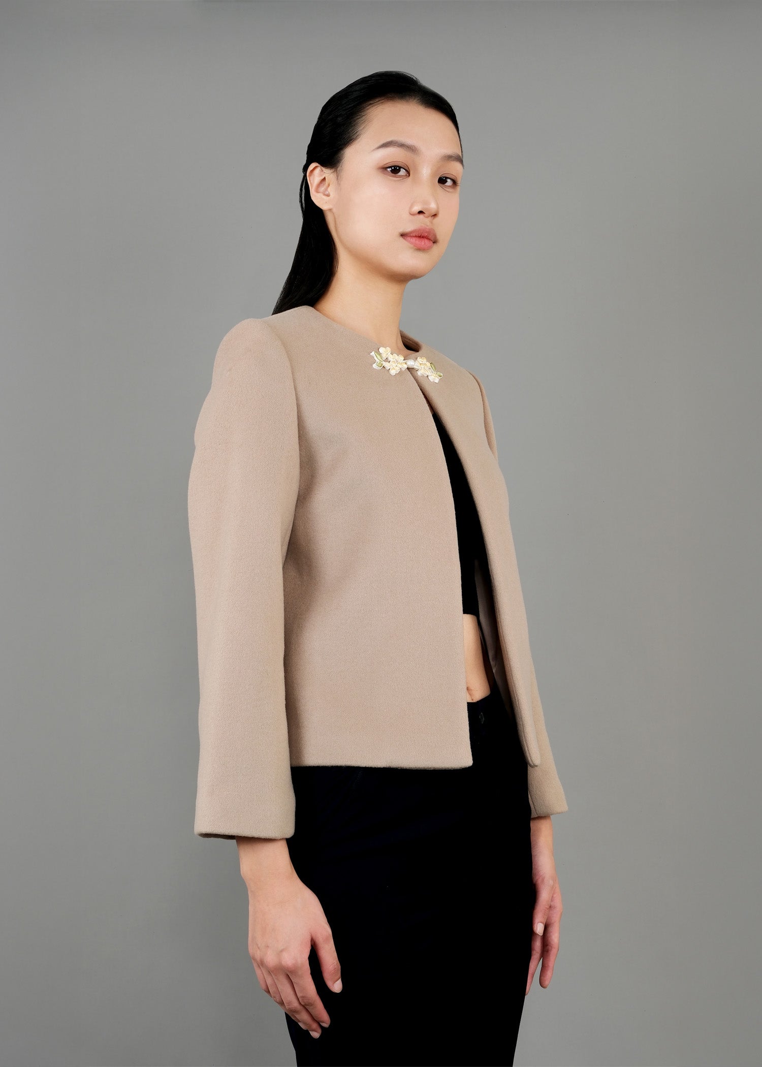 A woman stands in side view, wearing a stylish beige jacket with a round neckline and a decorative floral brooch at the front. The jacket has long sleeves and is paired with a black cropped top and dark trousers. 
一名女子站在側面，身穿時尚的米色圓領外套，胸前飾有花朵胸針。外套為長袖設計，內搭黑色露臍上衣和深色長褲。