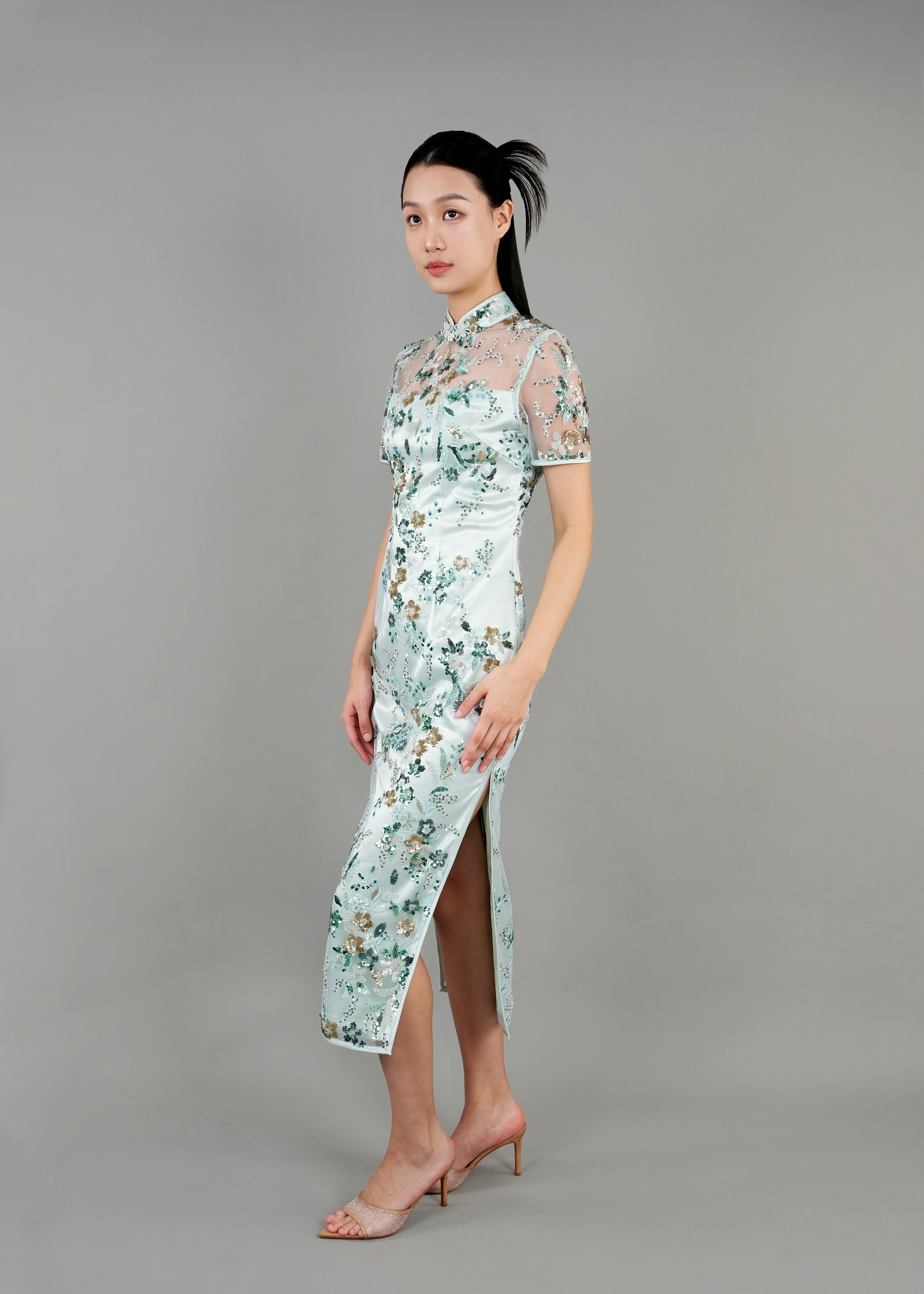 A woman stands in side view ,wearing a light blue cheongsam dress with floral patterns. The dress has a sheer, embroidered overlay and features a high collar and short sleeves. 
一名女子站在側面，身穿淺藍色花卉圖案旗袍。旗袍外罩一層薄紗刺繡，領口較高，袖子較短。