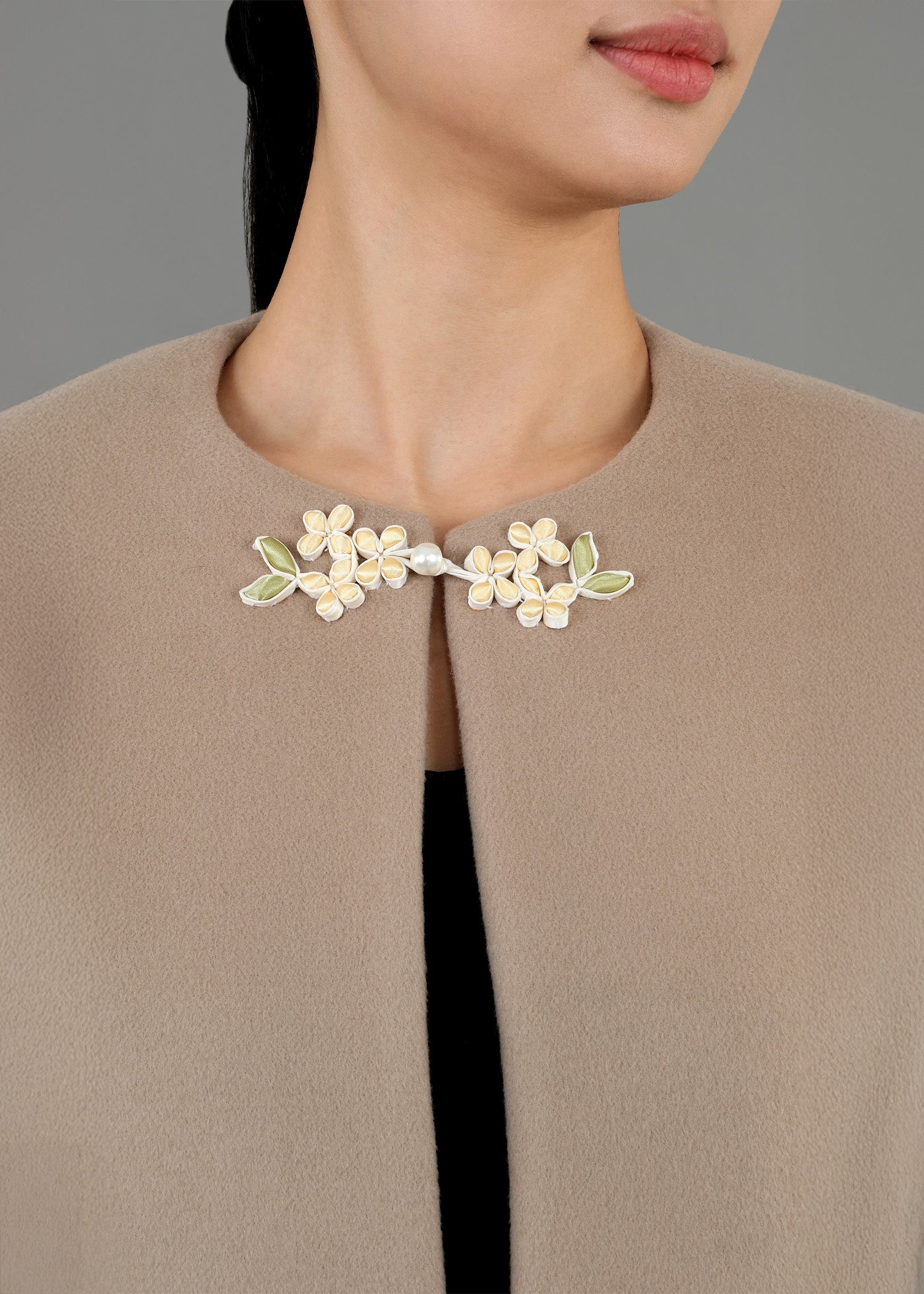 A close-up of a decorative floral brooch on a beige jacket. The brooch features delicate flowers in light yellow with green leaves, and a small pearl in the center, adding an elegant touch to the garment.
特寫鏡頭展現了一件米色外套上別著的一枚裝飾性花卉胸針。胸針上點綴著淡黃色的精緻花朵和綠色的葉子，中心鑲嵌著一顆小珍珠，為這件衣服增添了一抹優雅氣息。 
