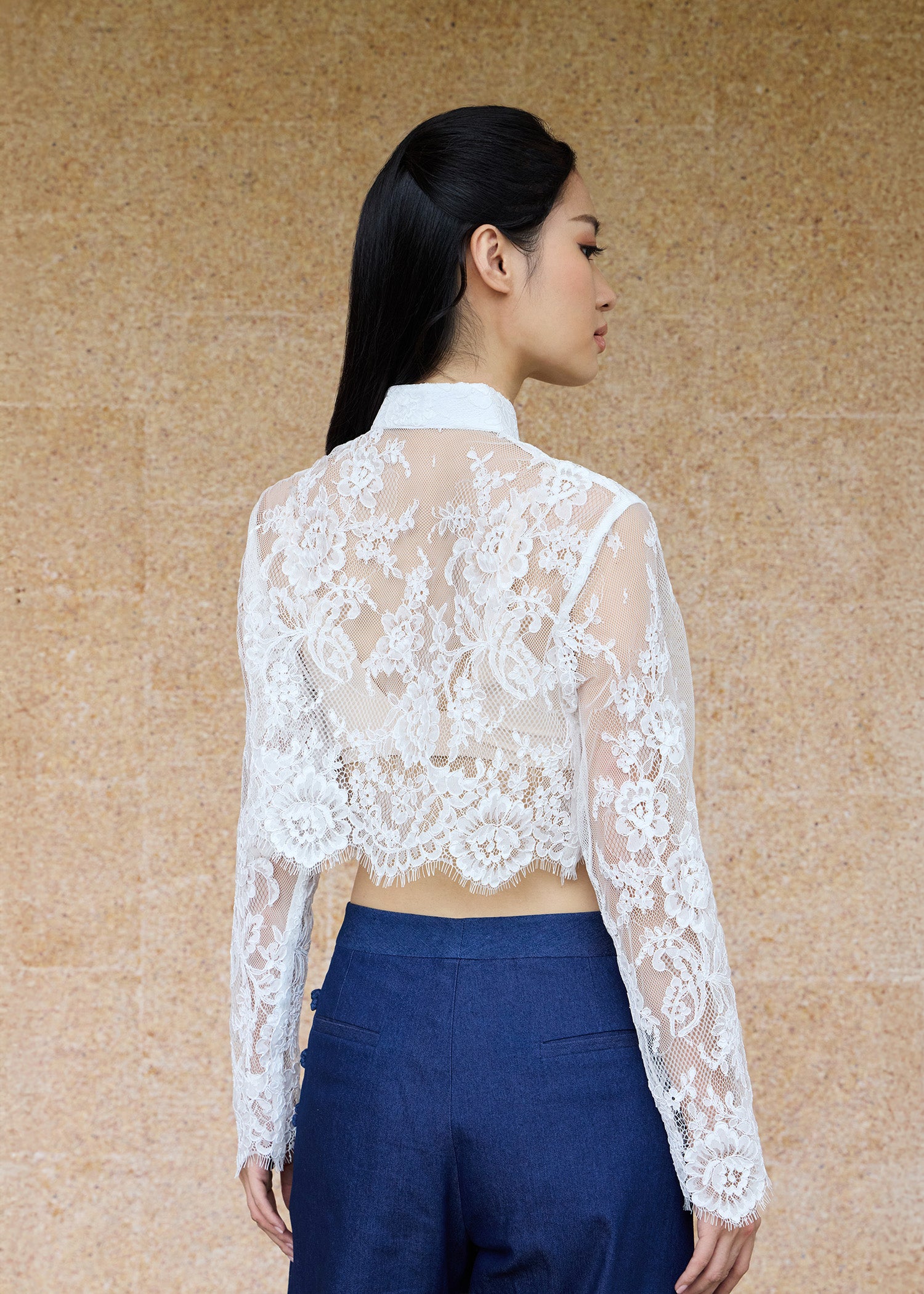Back view, half-body shot of an Asian woman wearing a long sleeve lace qipao jacket by Qipology, set against a yellow-beige background.