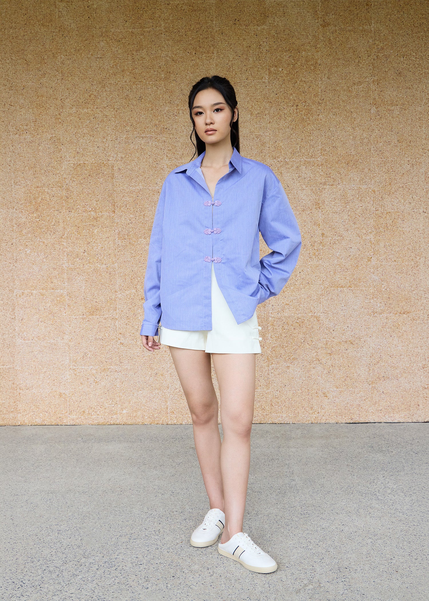 Front view, full-body shot of an Asian woman wearing a periwinkle blue oversize shirt and a white shorts by Qipology, set against a yellow-beige background.