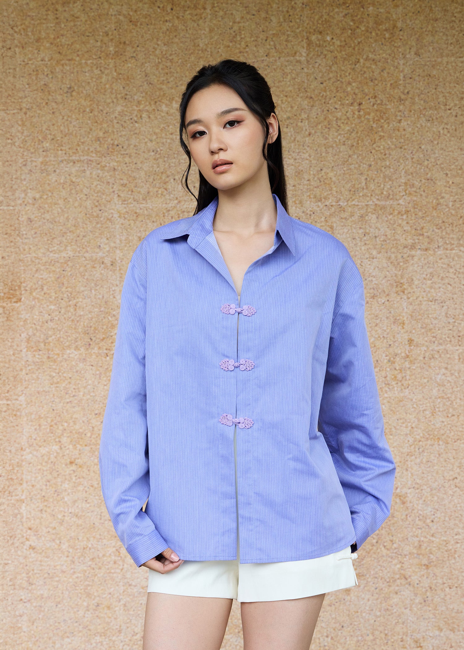 Front view, half-body shot of an Asian woman wearing a periwinkle blue oversize shirt and a white shorts by Qipology, set against a yellow-beige background.