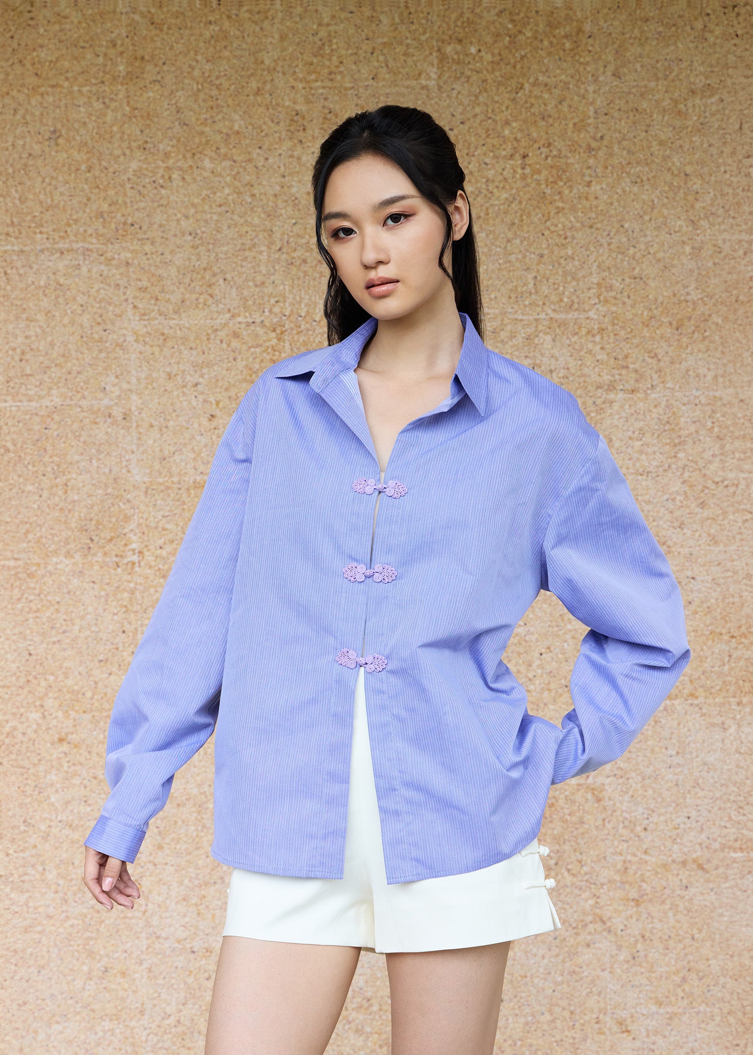 Front view, half-body shot of an Asian woman wearing a periwinkle blue oversize shirt and a white shorts by Qipology, set against a yellow-beige background.