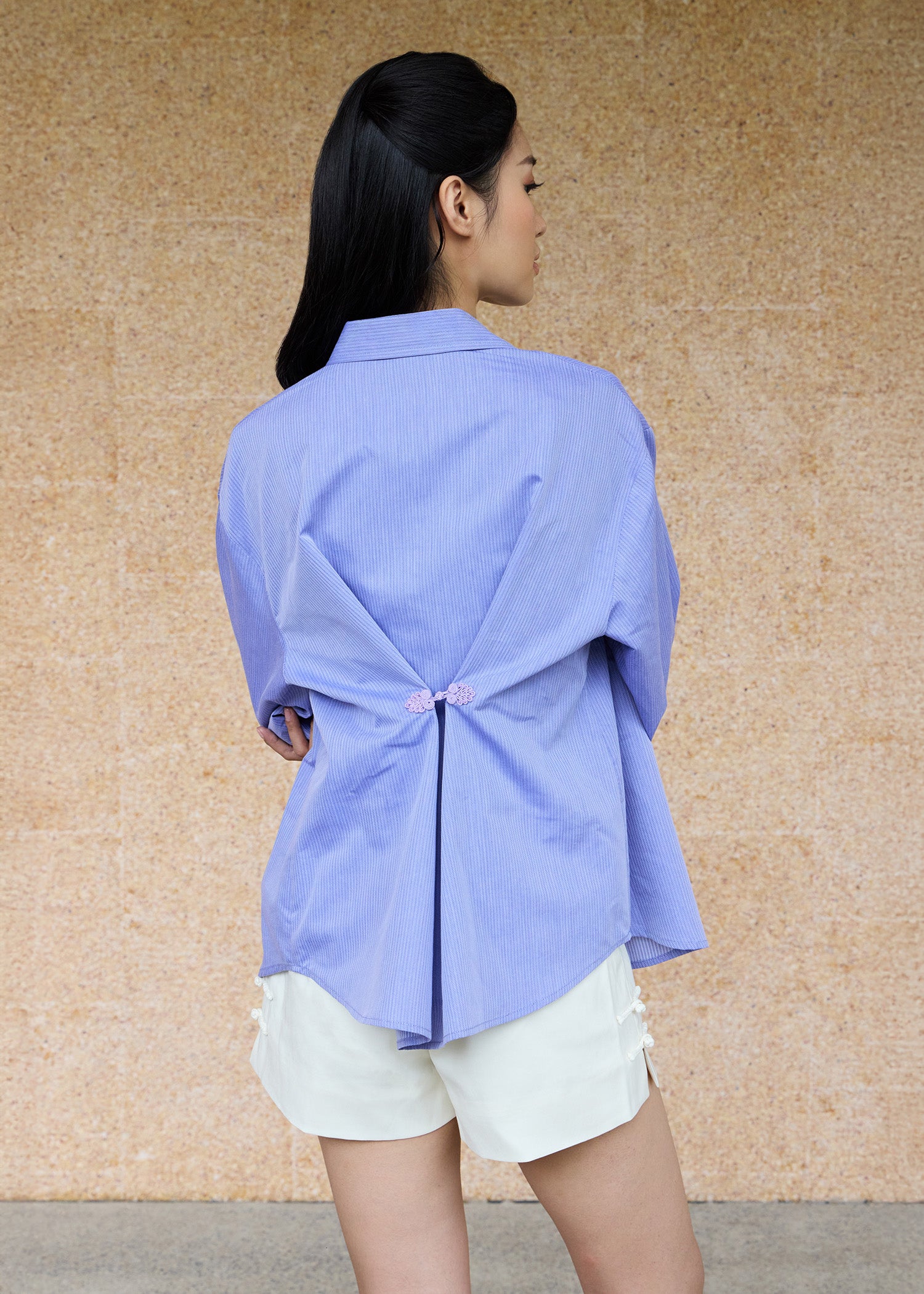 Back view, half-body shot of an Asian woman wearing a periwinkle blue oversize shirt and a white shorts by Qipology, set against a yellow-beige background.