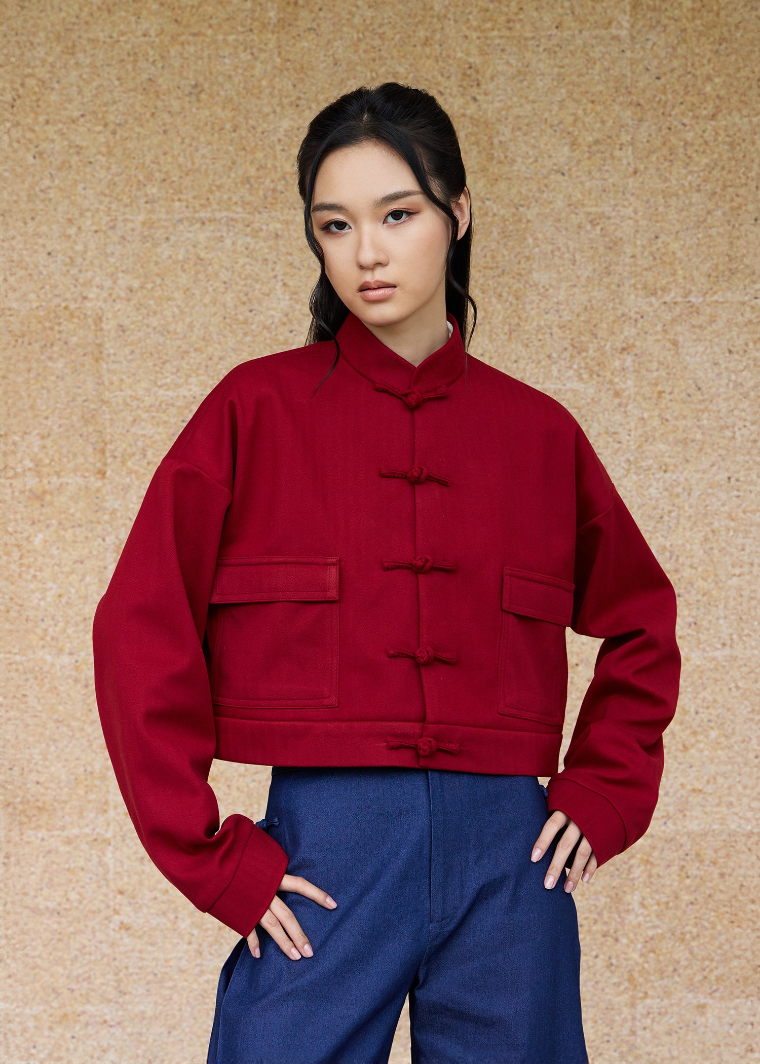 Front view, half-body shot of an Asian woman wearing a brick red Tang bomber by Qipology, set against a yellow-beige background.