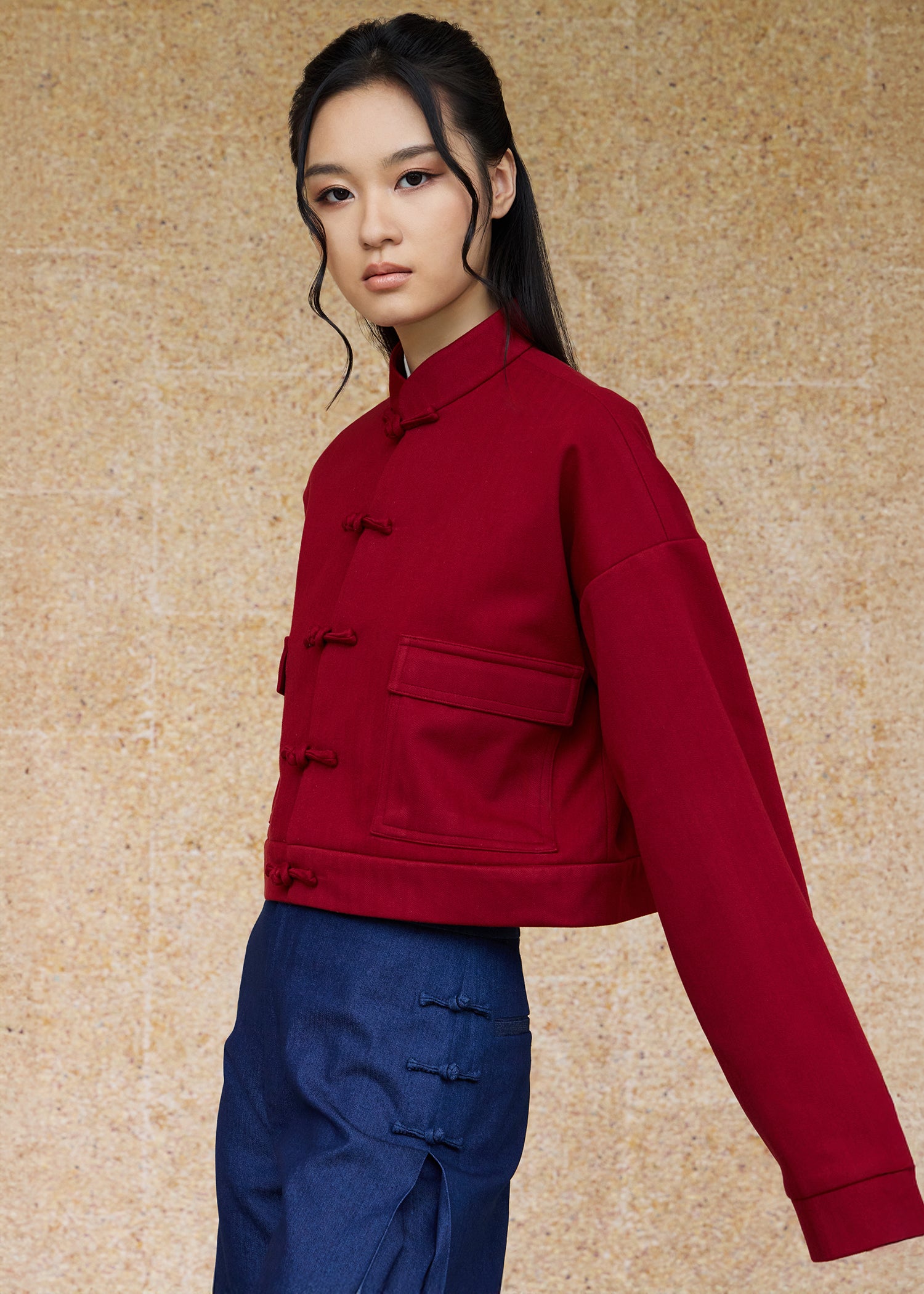 Front view, half-body shot of an Asian woman wearing a brick red Tang bomber by Qipology, set against a yellow-beige background.