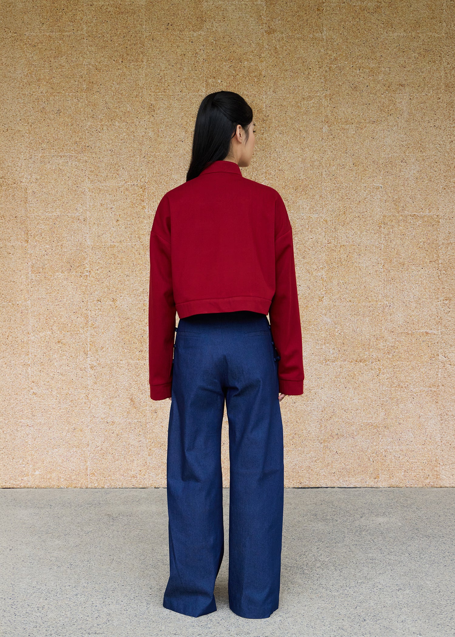 Back view, full-body shot of an Asian woman wearing a brick red Tang bomber by Qipology, set against a yellow-beige background.