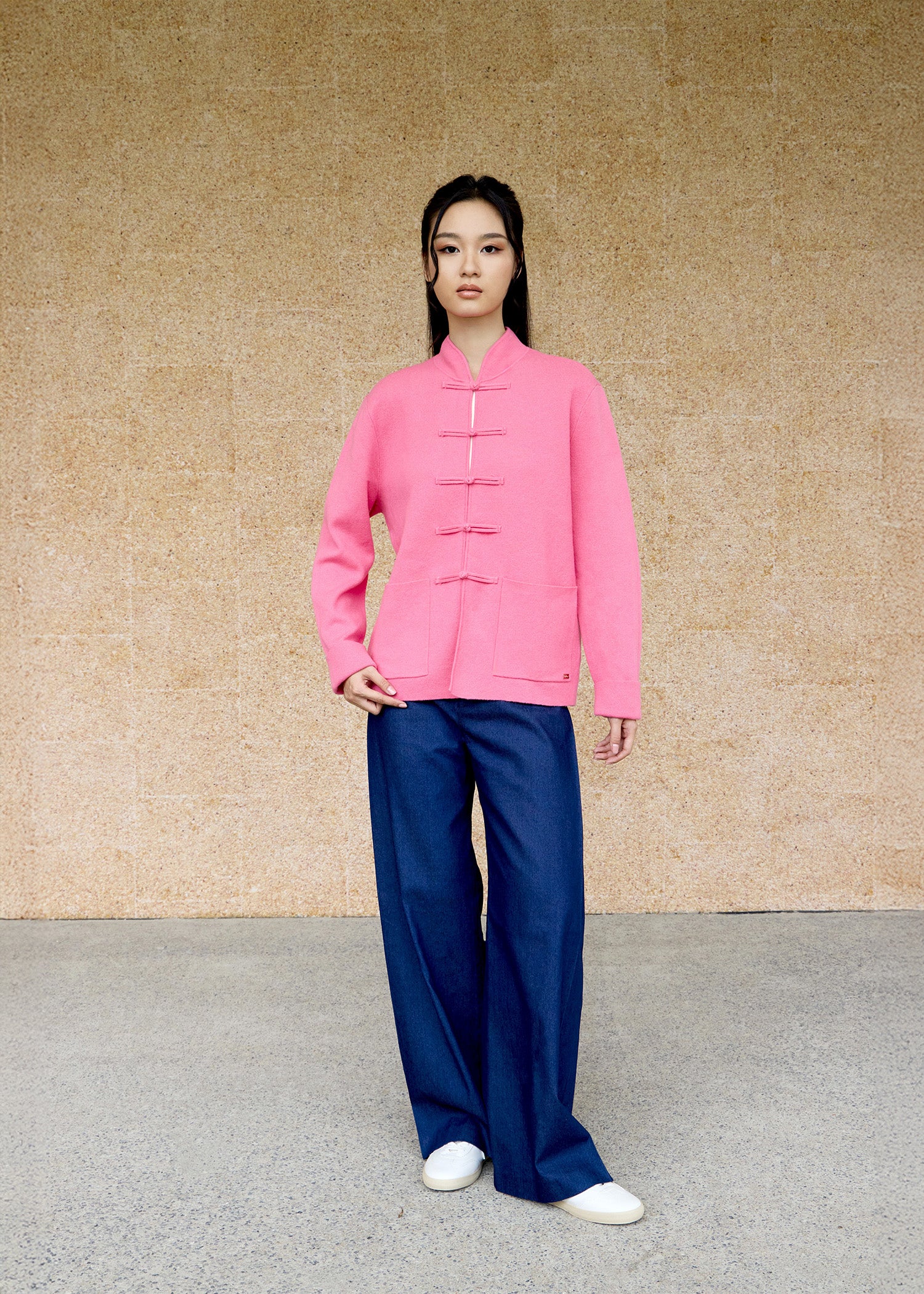 Front view, full-body shot of an Asian woman wearing a pink knitted Tang jacket by Qipology, set against a yellow-beige background.