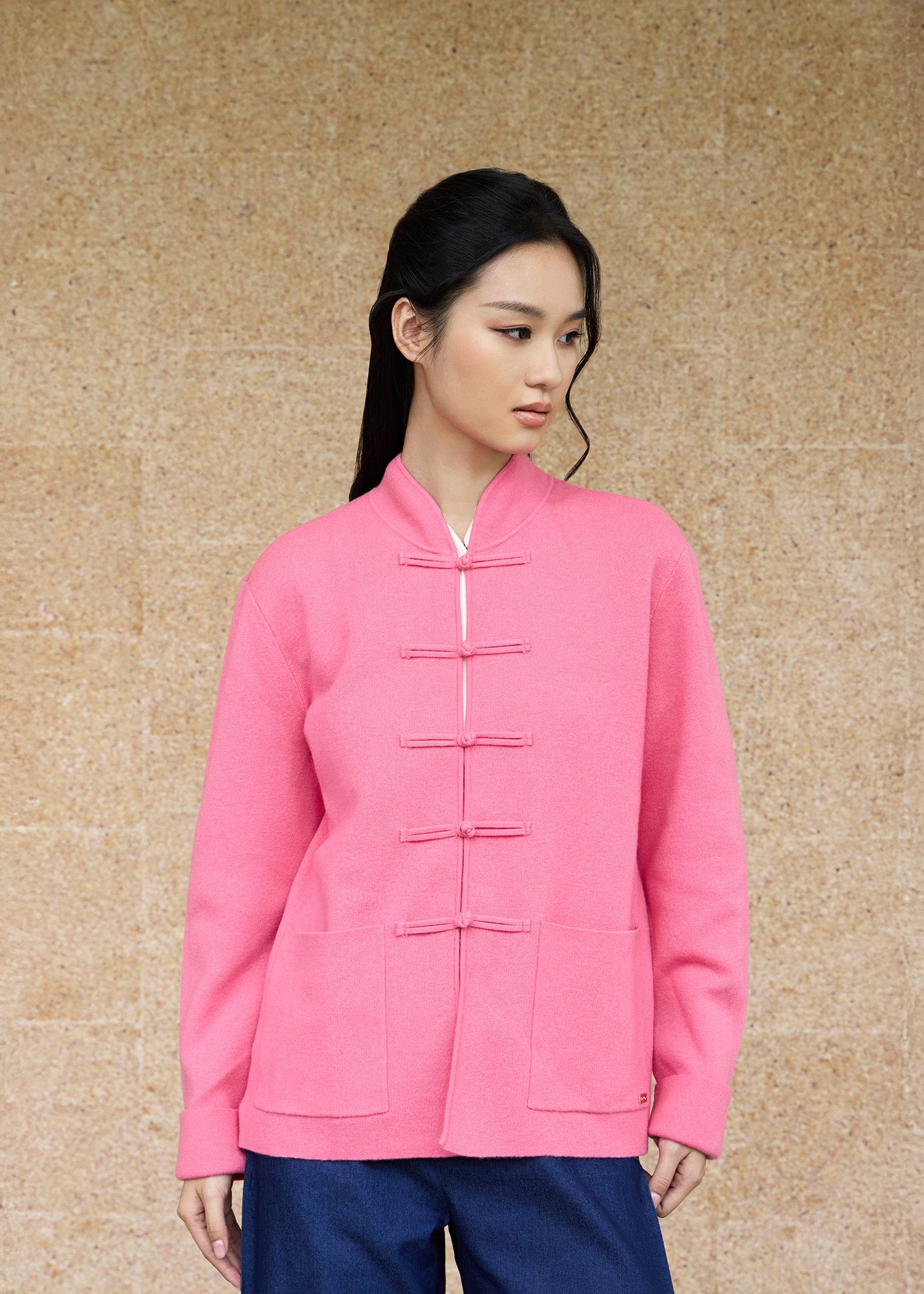 Front view, half-body shot of an Asian woman wearing a pink knitted Tang jacket by Qipology, set against a yellow-beige background.