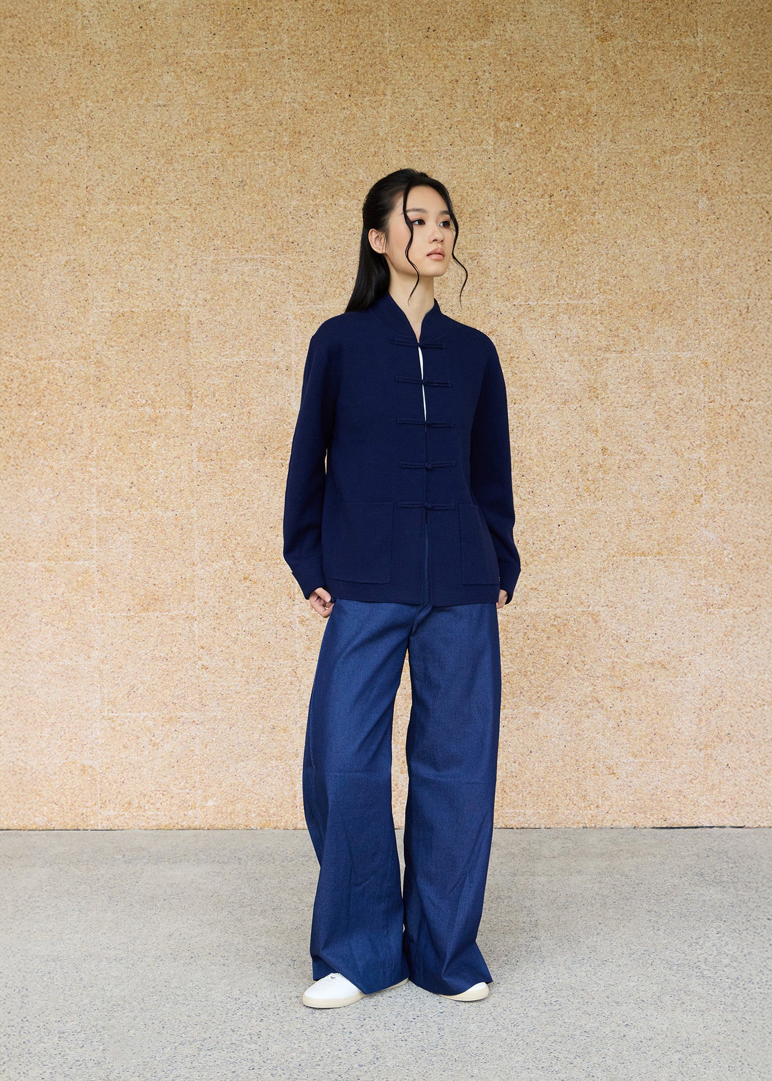 Front view, full-body shot of an Asian woman wearing a dark blue knitted Tang jacket over a qipao white top  and jeans byQipology, set against a yellow-beige background.