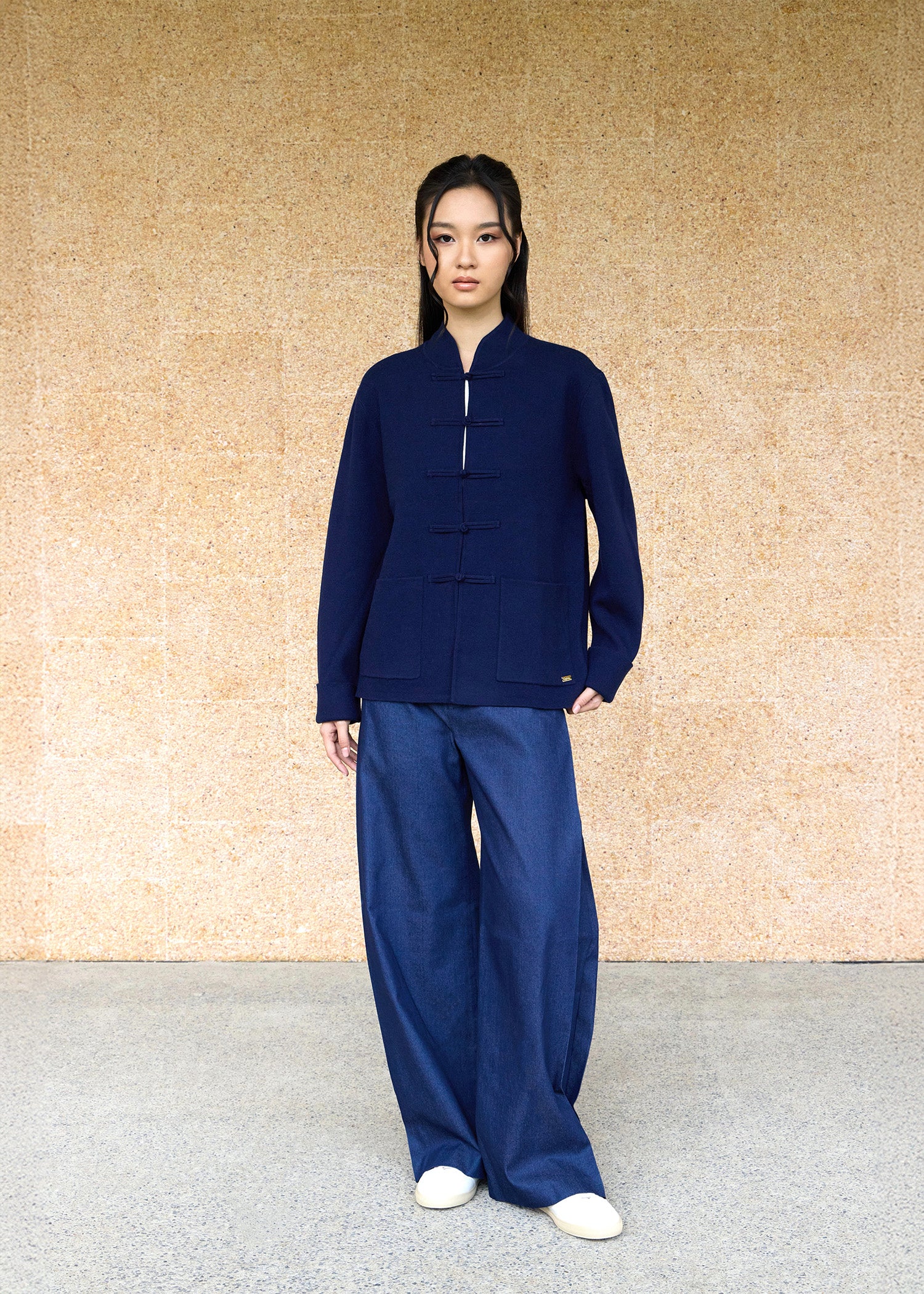 Front view, full body shot of an Asian woman wearing a dark blue knitted Tang jacket over a white qipao top and jeans by Qipology, set against a yellow-beige background.