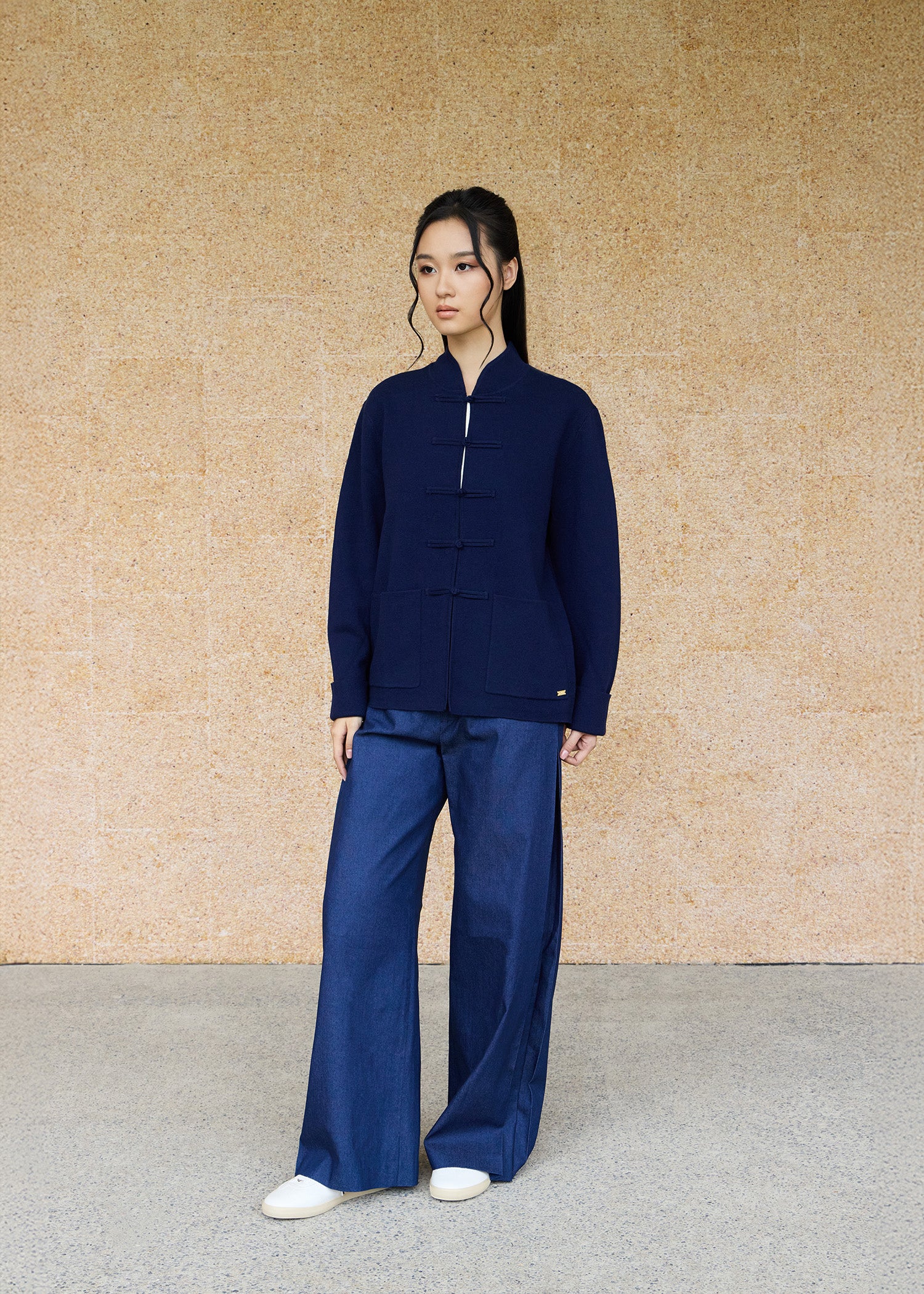 Front view, full body shot of an Asian woman wearing a dark blue knitted Tang jacket over a white qipao top and jeans by Qipology, set against a yellow-beige background.