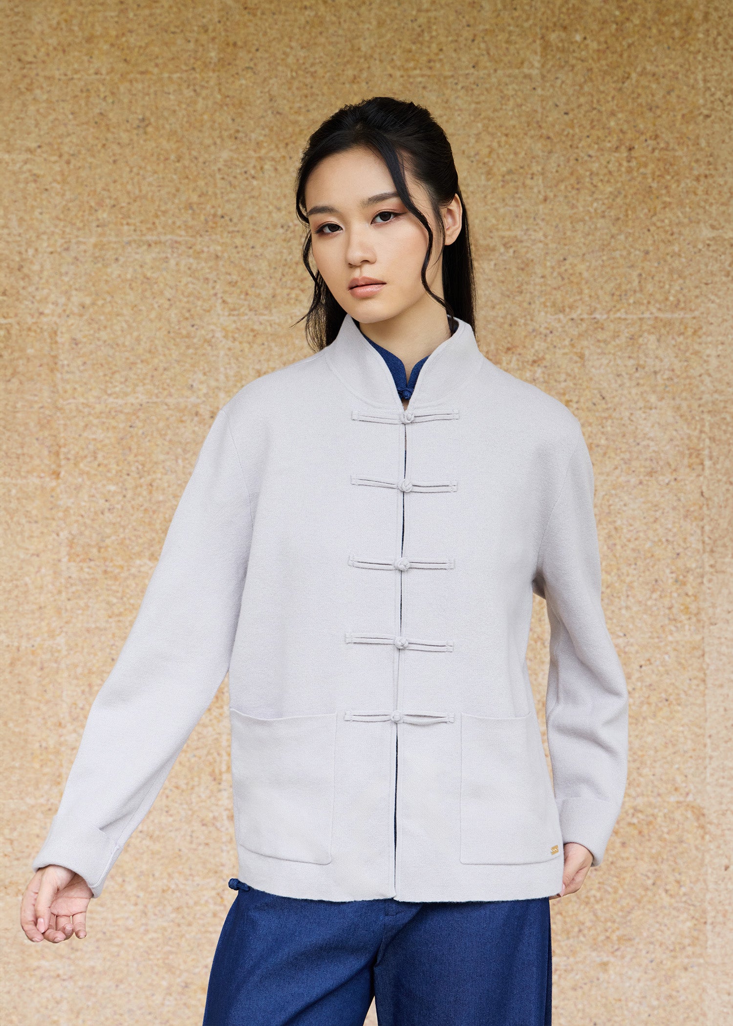 Front view, half-body shot of an Asian woman wearing a khaki grey knitted Tang jacket over a keyhole denim qipao top by Qipology, set against a yellow-beige background.