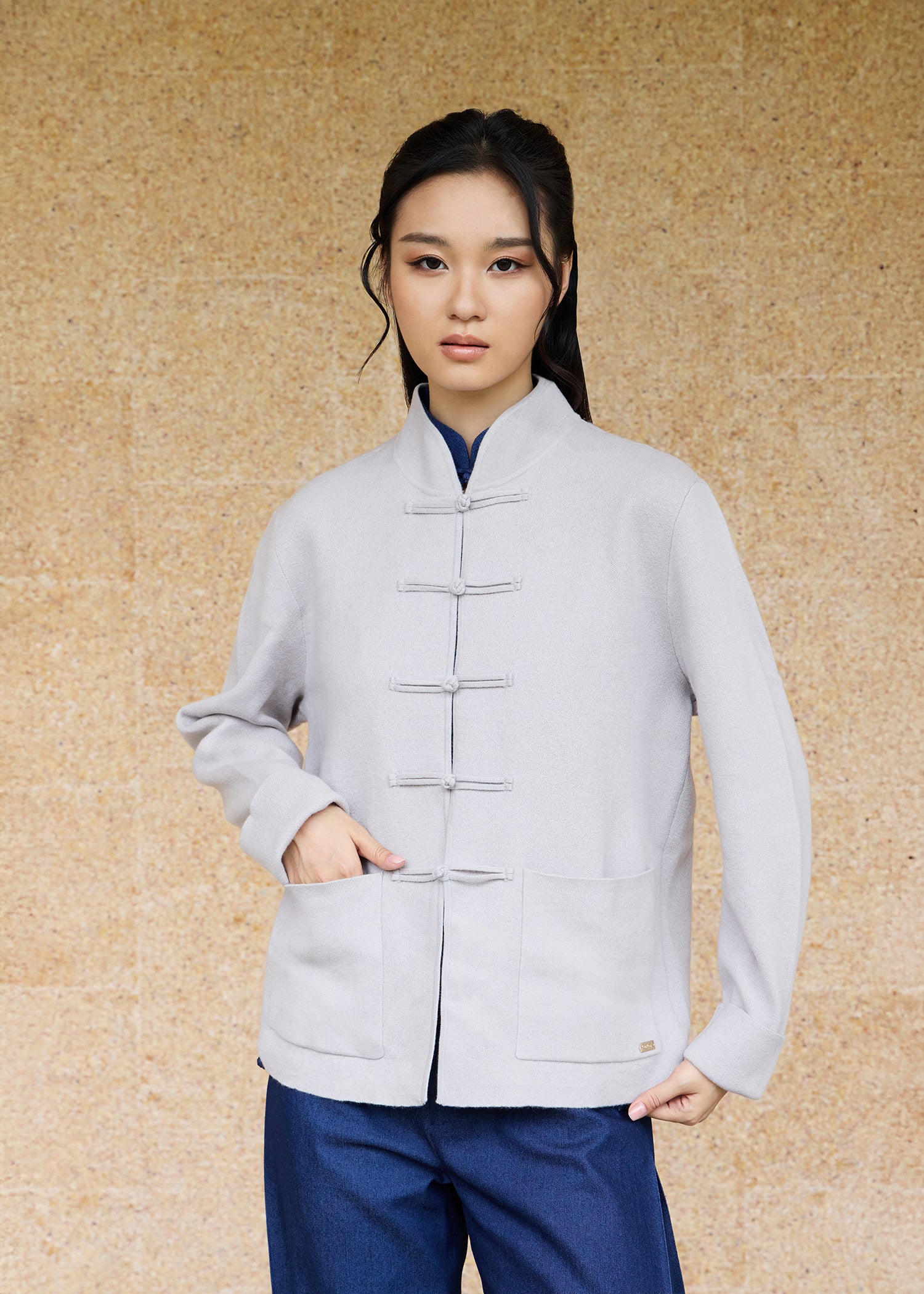 Front view, half-body shot of an Asian woman wearing a khaki grey knitted Tang jacket over a keyhole denim qipao top by Qipology, set against a yellow-beige background.