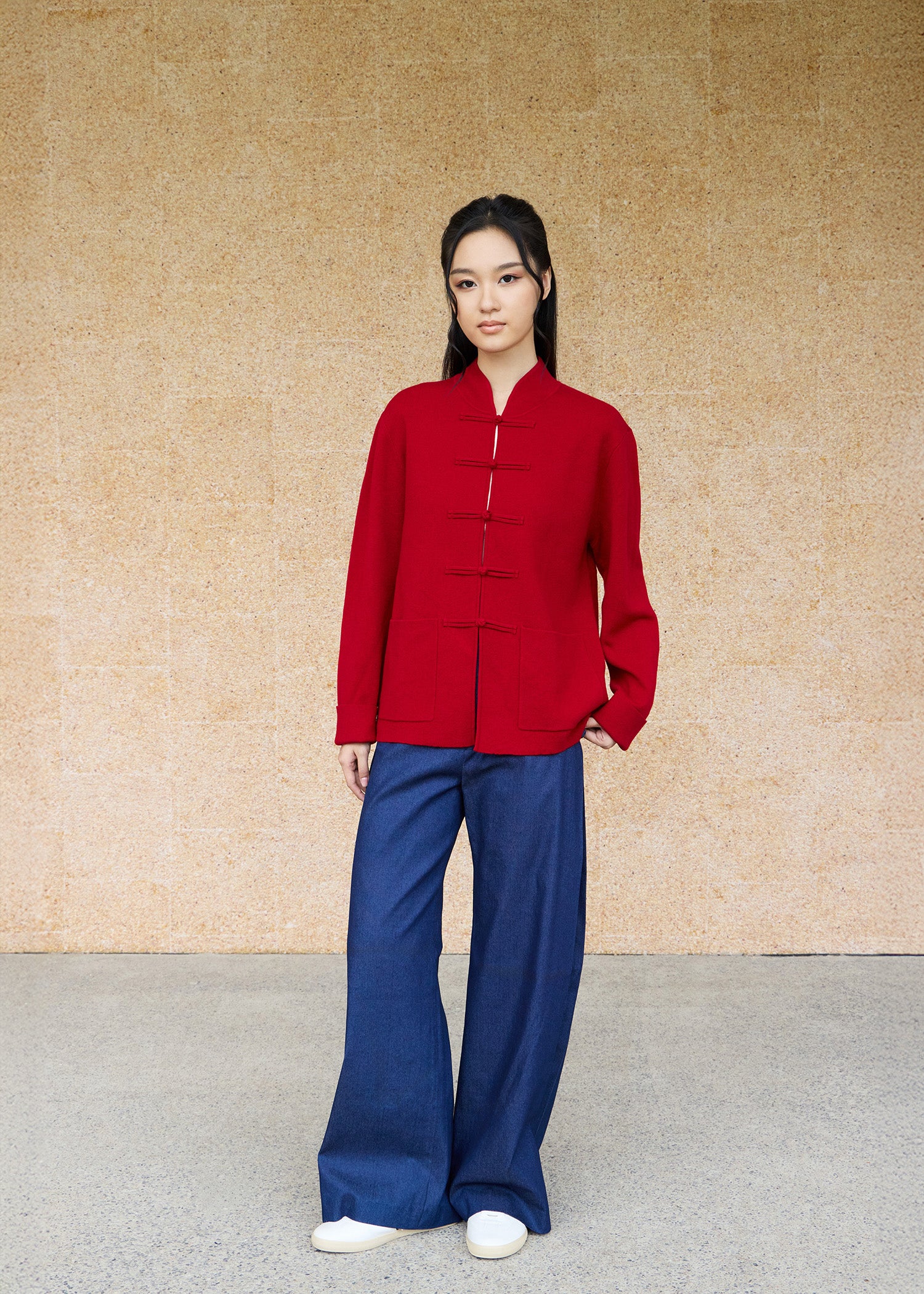 Front view, full-body shot of an Asian woman wearing a red knitted Tang jacket over top by Qipology, set against a yellow-beige background.