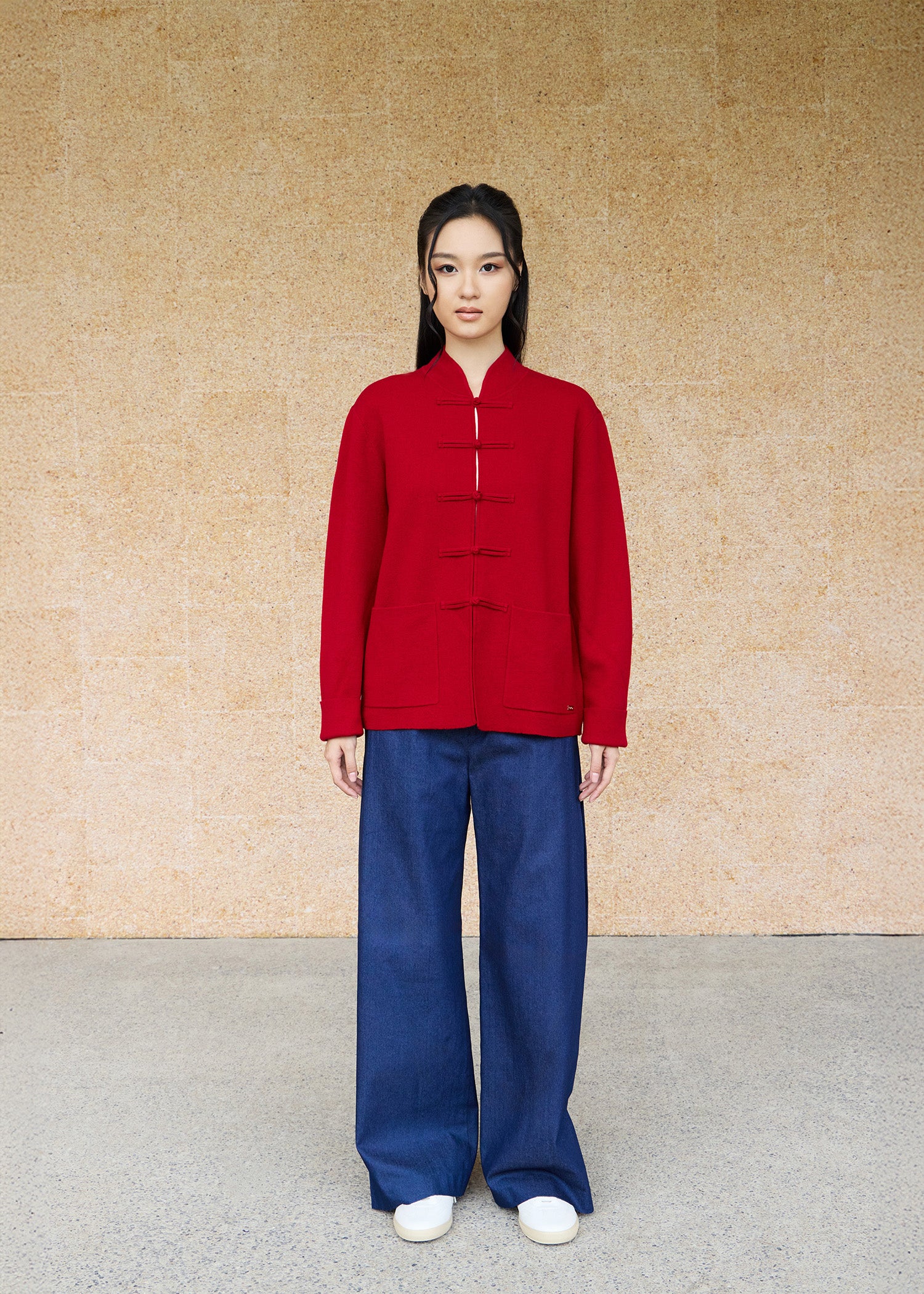 Front view, full-body shot of an Asian woman wearing a red knitted Tang jacket over top by Qipology, set against a yellow-beige background.