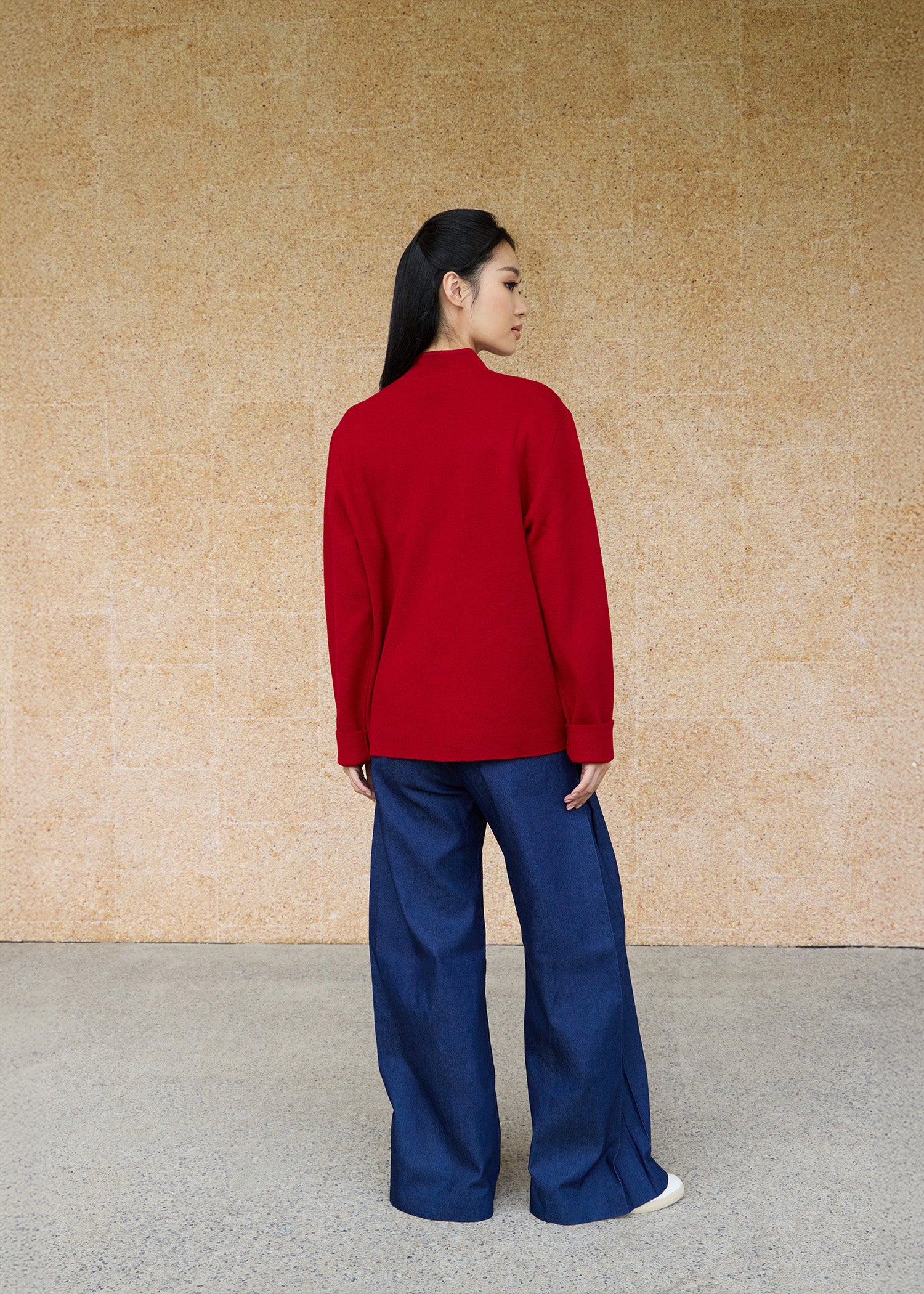 Back view, full-body shot of an Asian woman wearing a red knitted Tang jacket over top by Qipology, set against a yellow-beige background.