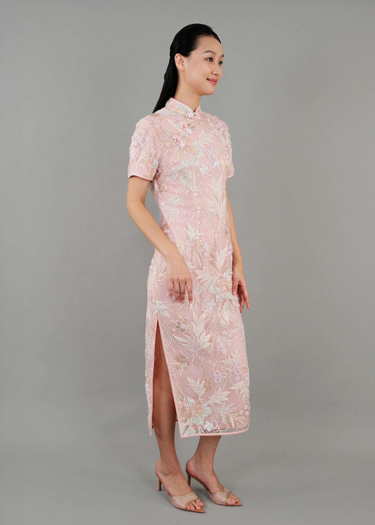 A woman stands in side view, wearing a light pink embroidered cheongsam dress. The dress features intricate floral patterns and a fitted silhouette with a high collar and short sleeves. 
一名女子站在側面，身穿淺粉色刺繡旗袍。旗袍飾有精緻的花卉圖案，修身剪裁，高領短袖。