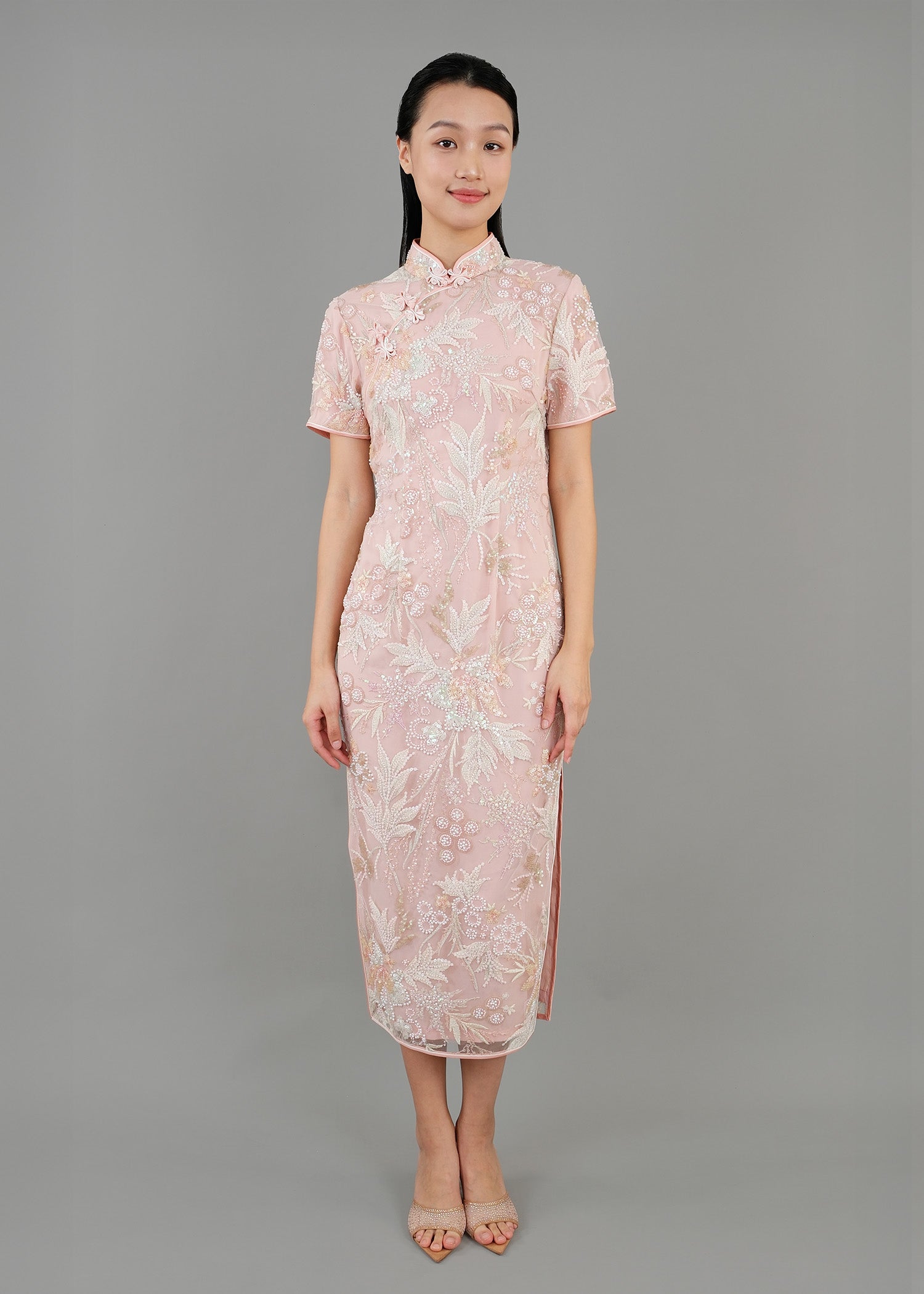 A woman stands in front view, wearing a light pink embroidered cheongsam dress. The dress features intricate floral patterns and a fitted silhouette with a high collar and short sleeves. 
一名女子站在正面，身穿淺粉色刺繡旗袍。旗袍飾有精緻的花卉圖案，修身剪裁，高領短袖。