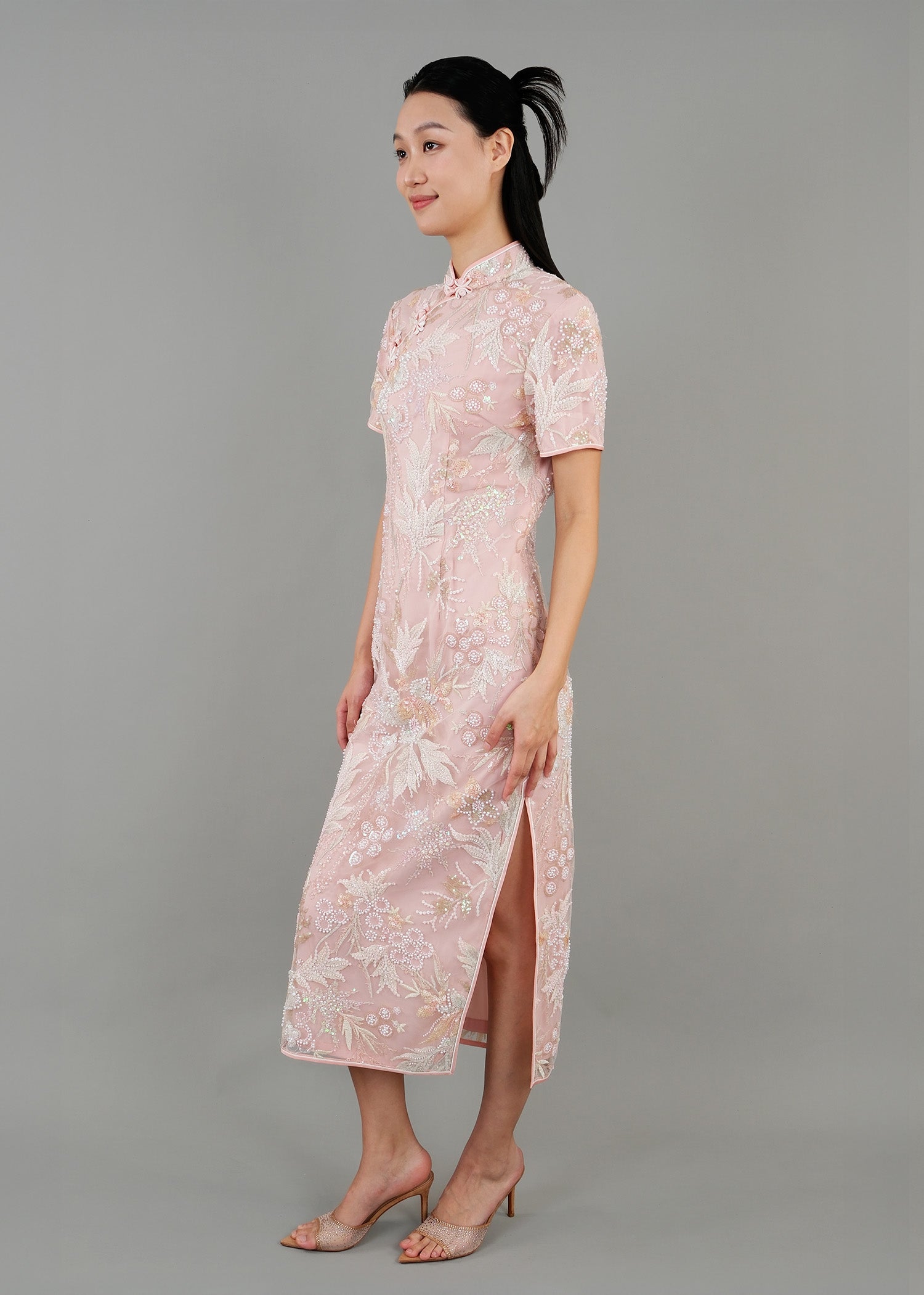 A woman stands in side view, wearing a light pink embroidered cheongsam dress. The dress features intricate floral patterns and a fitted silhouette with a high collar and short sleeves. 
一名女子站在側面，身穿淺粉色刺繡旗袍。旗袍飾有精緻的花卉圖案，修身剪裁，高領短袖。