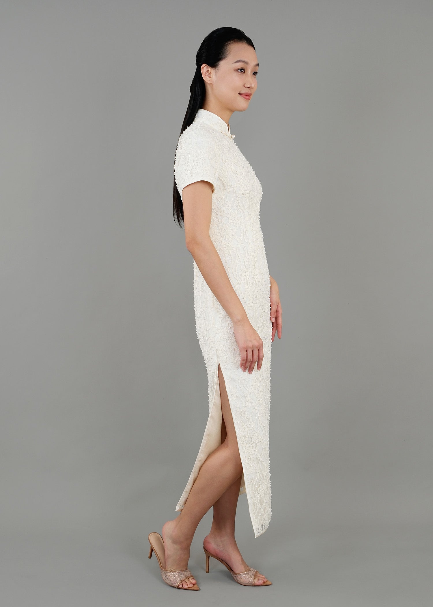 A woman stands in side view, wearing a white high-neck dress with short sleeves and textured detailing, featuring a side slit.
一名女子站在側面，身穿白色高領短袖連身裙，帶有紋理細節，側面有開衩。