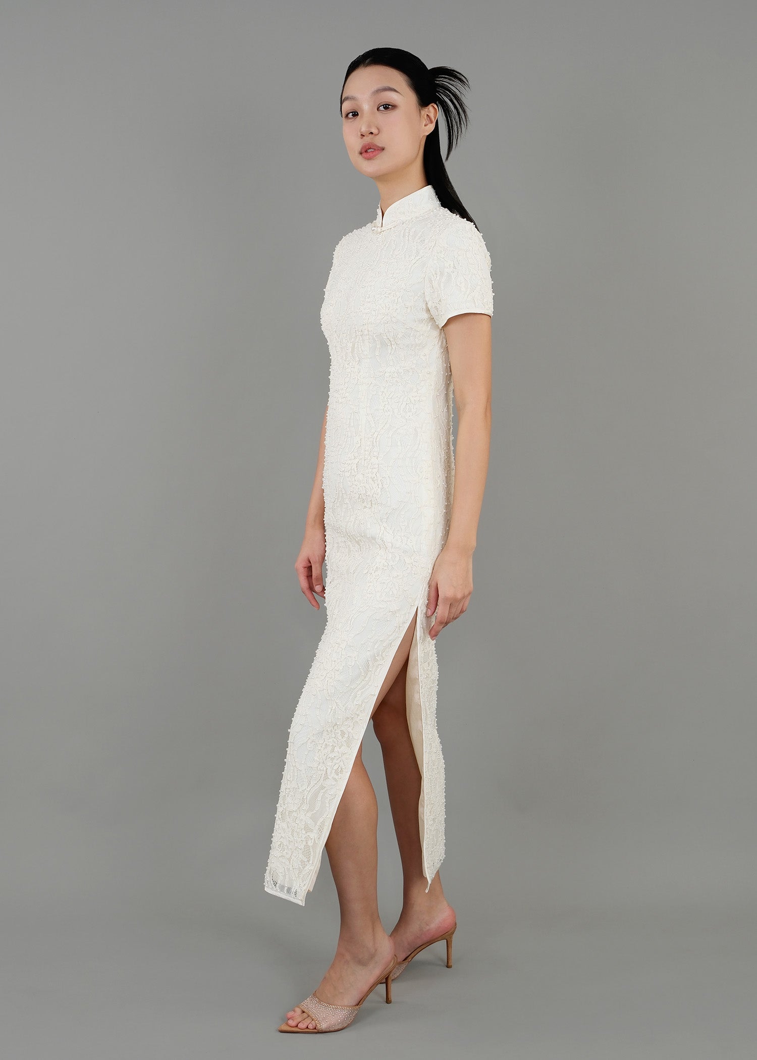 A woman stands in side view, wearing a white high-neck dress with short sleeves and textured detailing, featuring a side slit.
一名女子站在側面，身穿白色高領短袖連身裙，帶有紋理細節，側面有開衩。