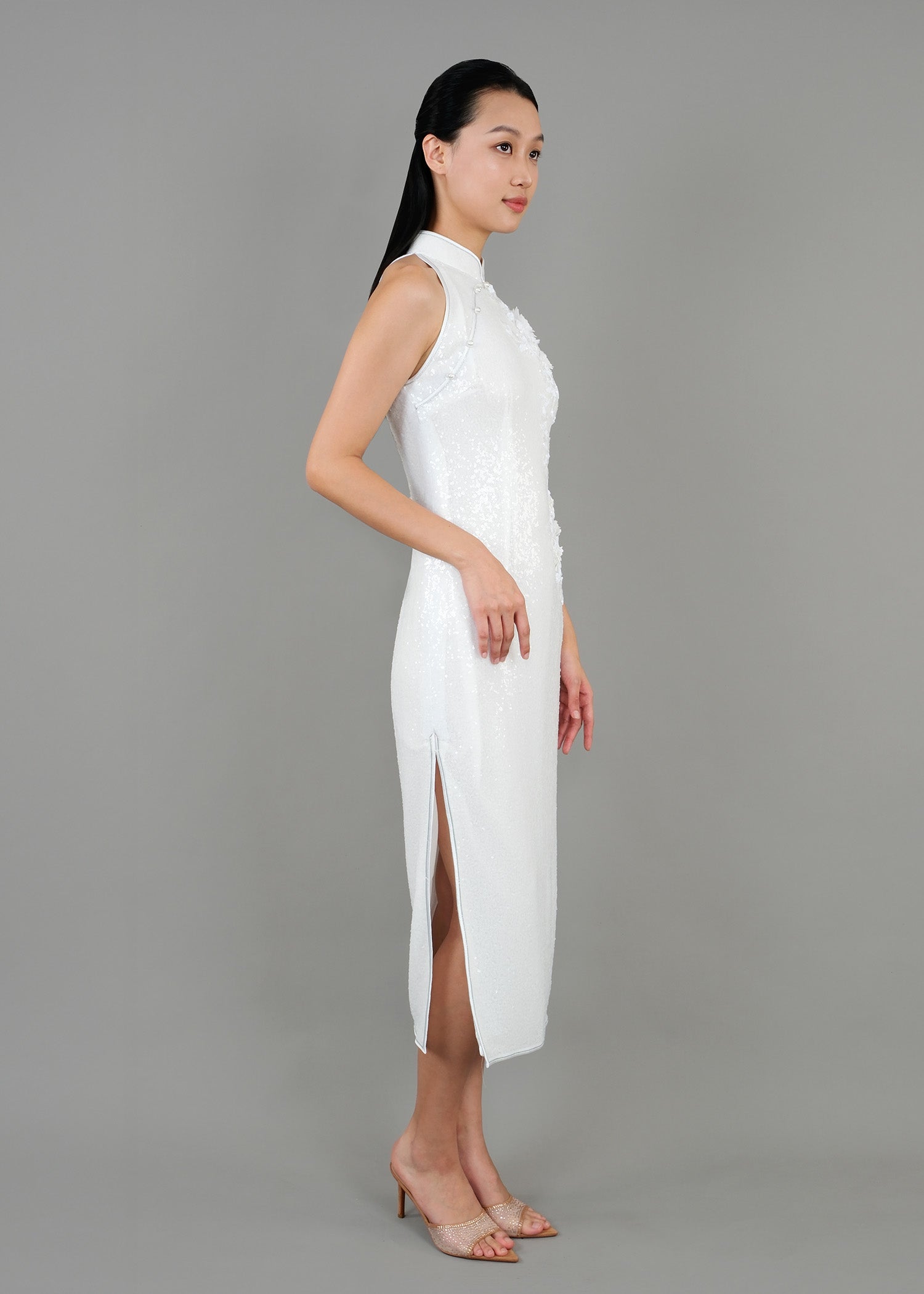 A woman is shown in side view, wearing a white high-neck dress with delicate floral patterns. The dress has a fitted silhouette that falls to the knee, featuring a side slit for added elegance.
一位女子身穿白色高領連身裙，飾有精緻的花卉圖案。洋裝剪裁修身，長度及膝蓋，側開衩更顯優雅。