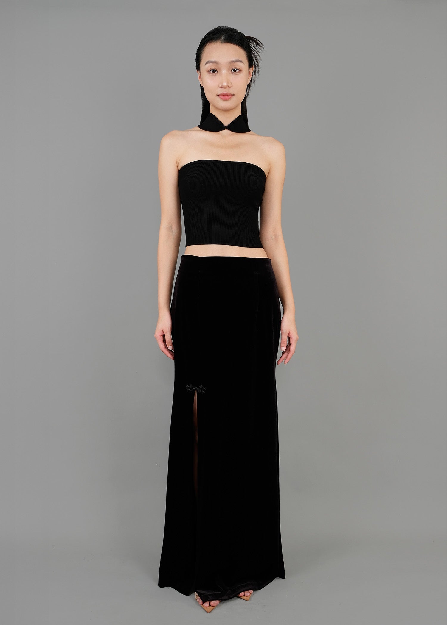 A woman stands in front view, wearing a stylish black outfit. The outfit consists of a strapless cropped top that fits snugly and a long black skirt with a slit on the left side. The skirt features a floral detail near the slit.
一名女子站在正面，身穿一套時尚的黑色套裝。這套服裝包括一件貼身的露肩短上衣和一條左側開衩的黑色長裙。裙子開衩附近飾有花朵圖案。