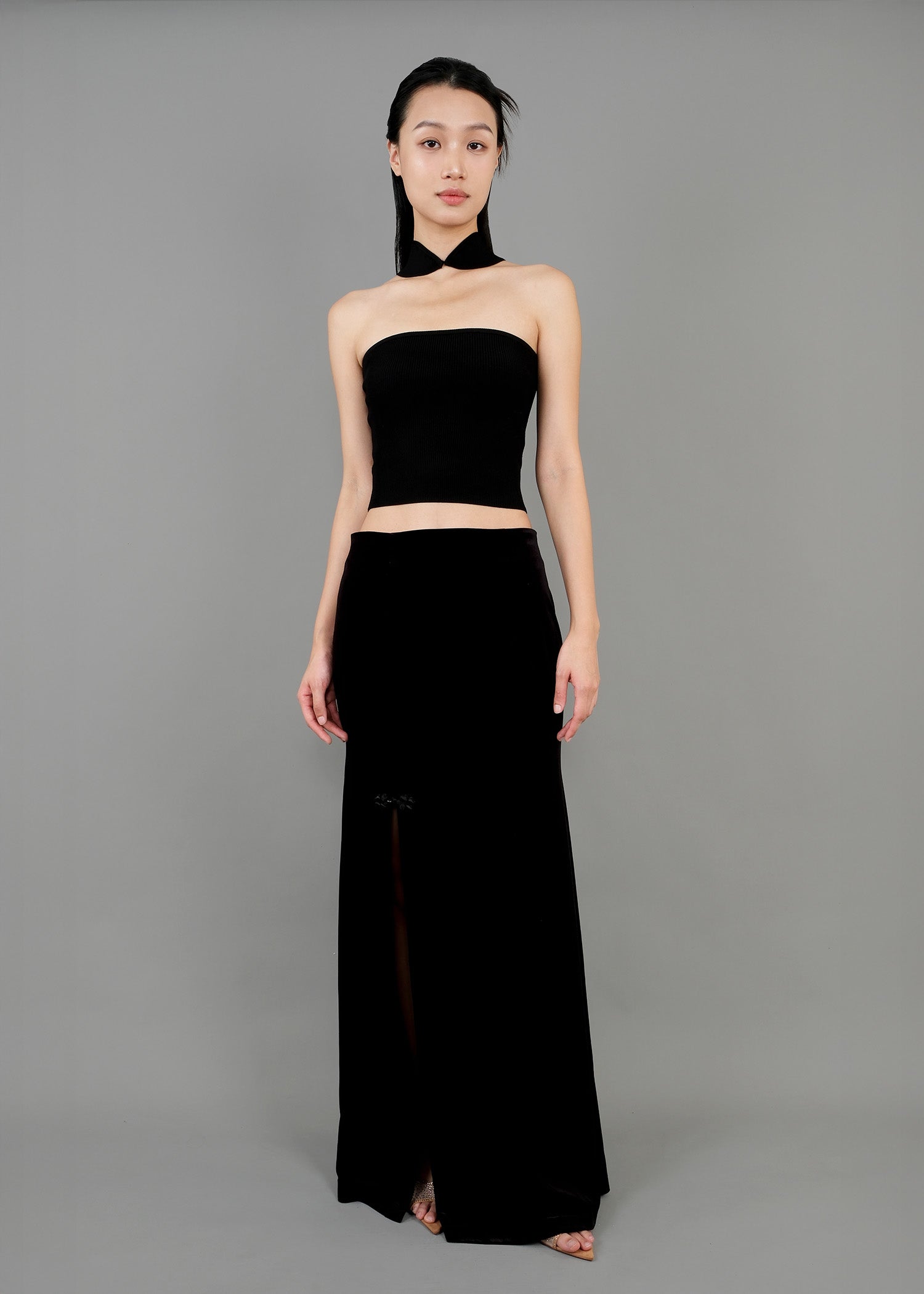 A woman stands in front view, wearing a stylish black outfit. The outfit consists of a strapless cropped top that fits snugly and a long black skirt with a slit on the left side. The skirt features a floral detail near the slit.
一名女子站在正面，身穿一套時尚的黑色套裝。這套服裝包括一件貼身的露肩短上衣和一條左側開衩的黑色長裙。裙子開衩附近飾有花朵圖案。
