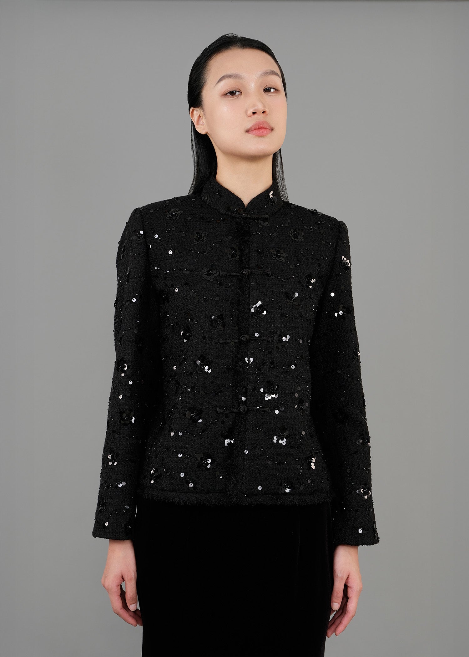 A woman stands in front view, wearing a black sequined jacket with a high collar and long sleeves, paired with a long black velvet skirt that features a slit. 
一名女子站在正面，身穿黑色亮片高領長袖外套，搭配黑色開衩長天鵝絨裙。