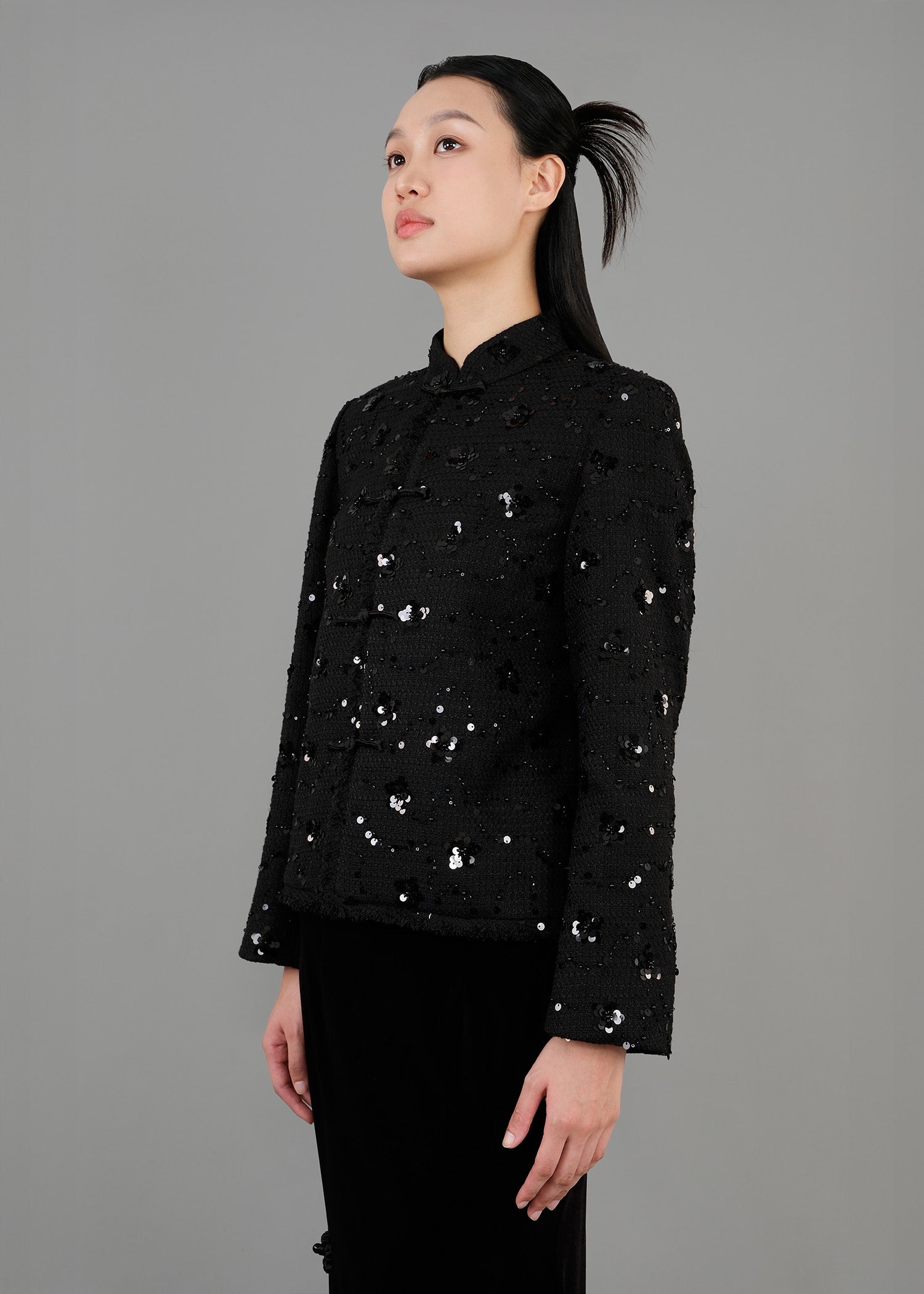 A woman stands in side view, wearing a black sequined jacket with a high collar and long sleeves, paired with a long black velvet skirt that features a slit. 
一名女子站在側面，身穿黑色亮片高領長袖外套，搭配黑色開衩長天鵝絨裙。