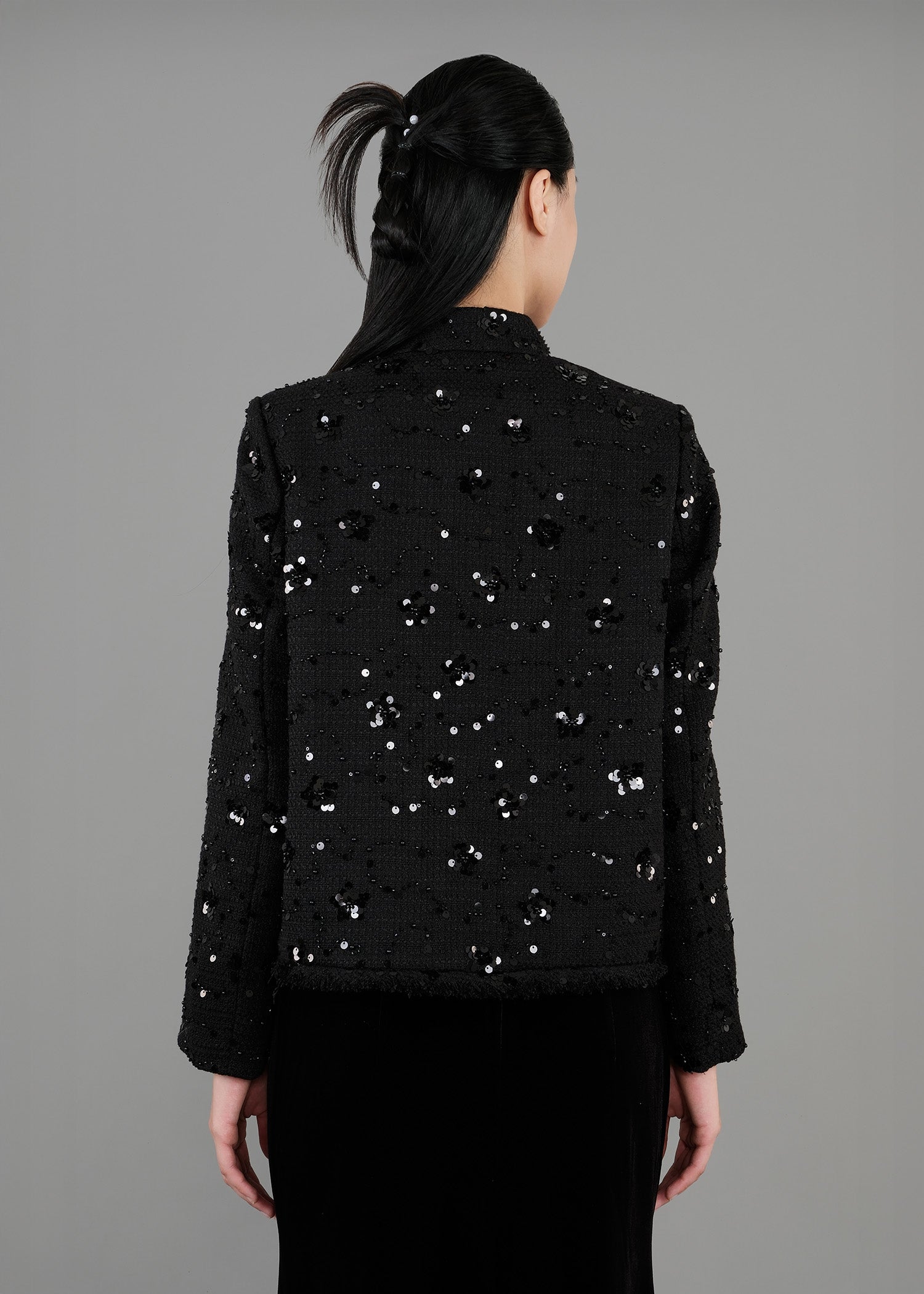 A woman stands in back view,  wearing a black sequined jacket with a high collar and long sleeves, paired with a long black velvet skirt that features a slit.
一名女子站在背影中，身穿黑色亮片高領長袖外套，搭配黑色開衩長天鵝絨裙。