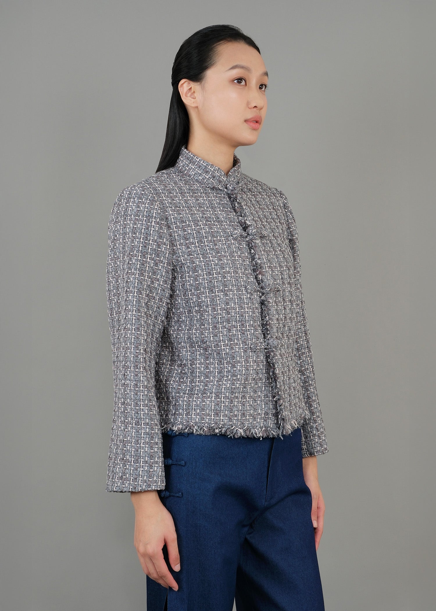A woman shown in side view, wearing a textured green gray button-up top with a high collar and loose blue pants, exuding a modern and sophisticated style.
一名女子站在側邊，身穿綠灰色高領質感紐扣上衣，搭配寬鬆藍色褲子，展現出現代精緻的風格。