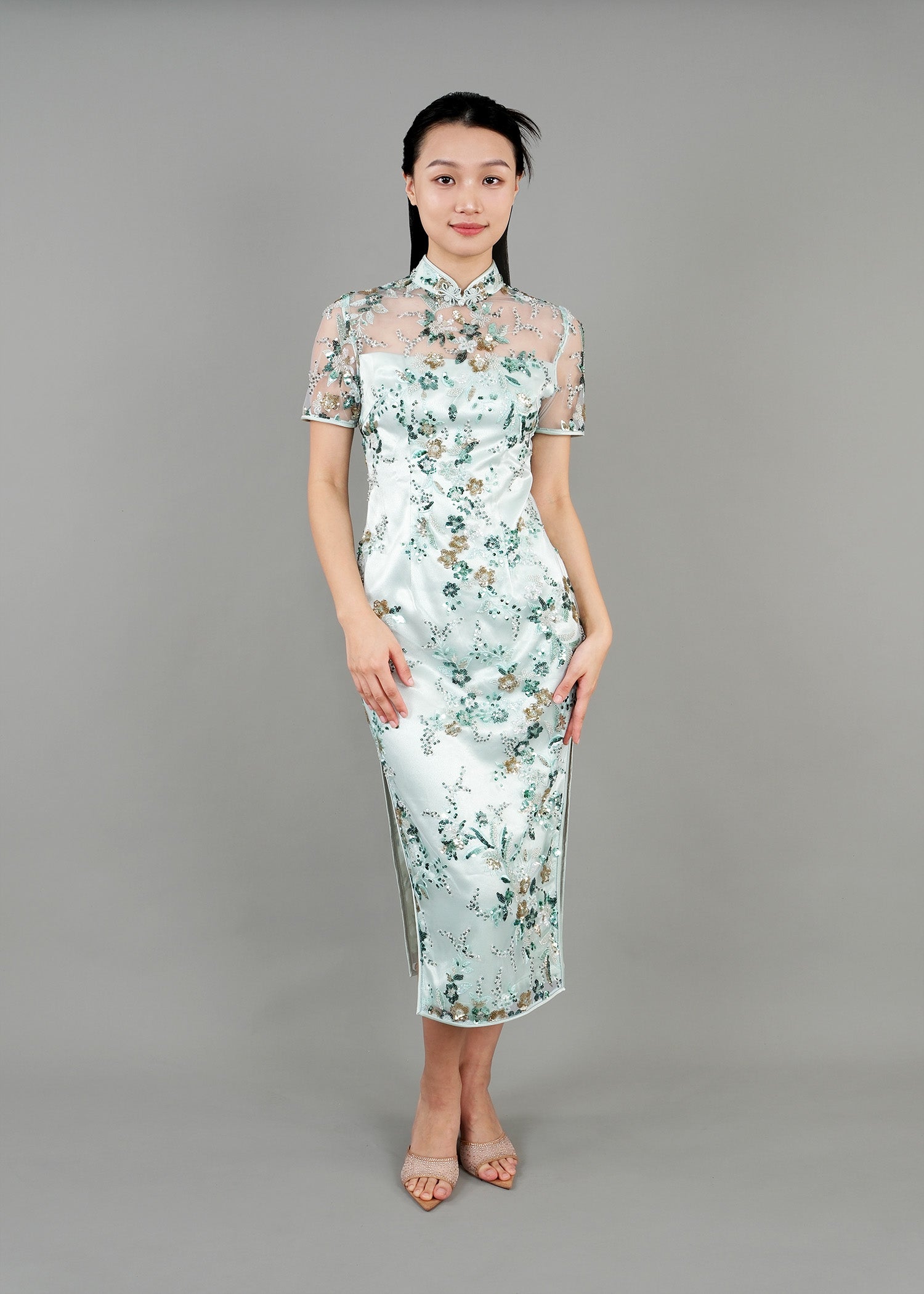 A woman stands in front view ,wearing a light blue cheongsam dress with floral patterns. The dress has a sheer, embroidered overlay and features a high collar and short sleeves. 
一名女子站在正面，身穿淺藍色花卉圖案旗袍。旗袍外罩一層薄紗刺繡，領口較高，袖子較短。
